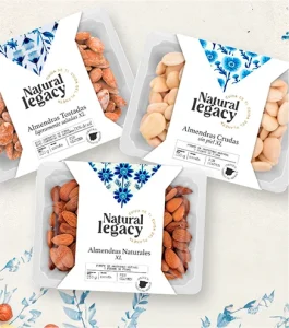 Tres paquetes de nueces Natural Legacy sobre un fondo floral. Los paquetes incluyen almendras XL, almendras crudas sin piel y almendras tostadas con sal del Himalaya. Cada uno tiene un diseño blanco y azul con secciones transparentes que muestran las nueces en su interior.