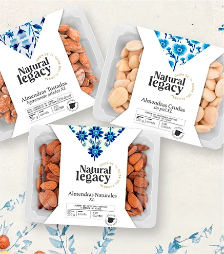 Tres paquetes de nueces Natural Legacy sobre un fondo floral. Los paquetes incluyen almendras XL, almendras crudas sin piel y almendras tostadas con sal del Himalaya. Cada uno tiene un diseño blanco y azul con secciones transparentes que muestran las nueces en su interior.