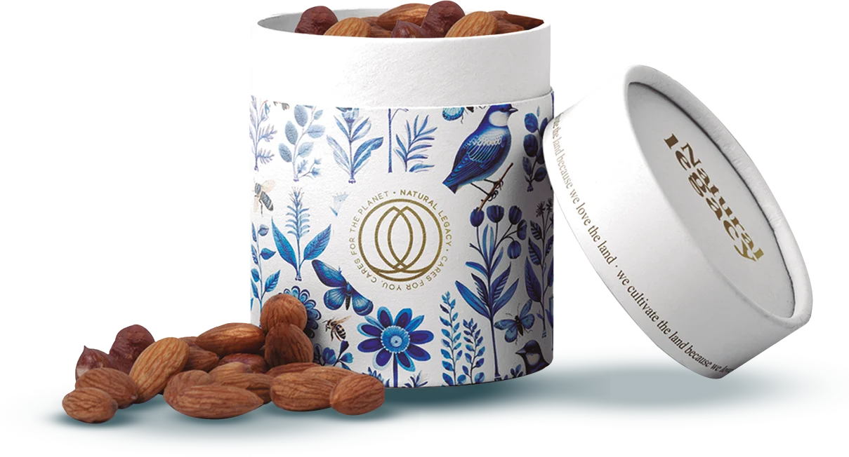 Un recipiente cilíndrico con un diseño floral y de pájaros en color azul está abierto y en su interior se ven almendras. La tapa, que se encuentra junto al recipiente, presenta el logotipo dorado y un mensaje de sostenibilidad. Hay algunas almendras esparcidas cerca.